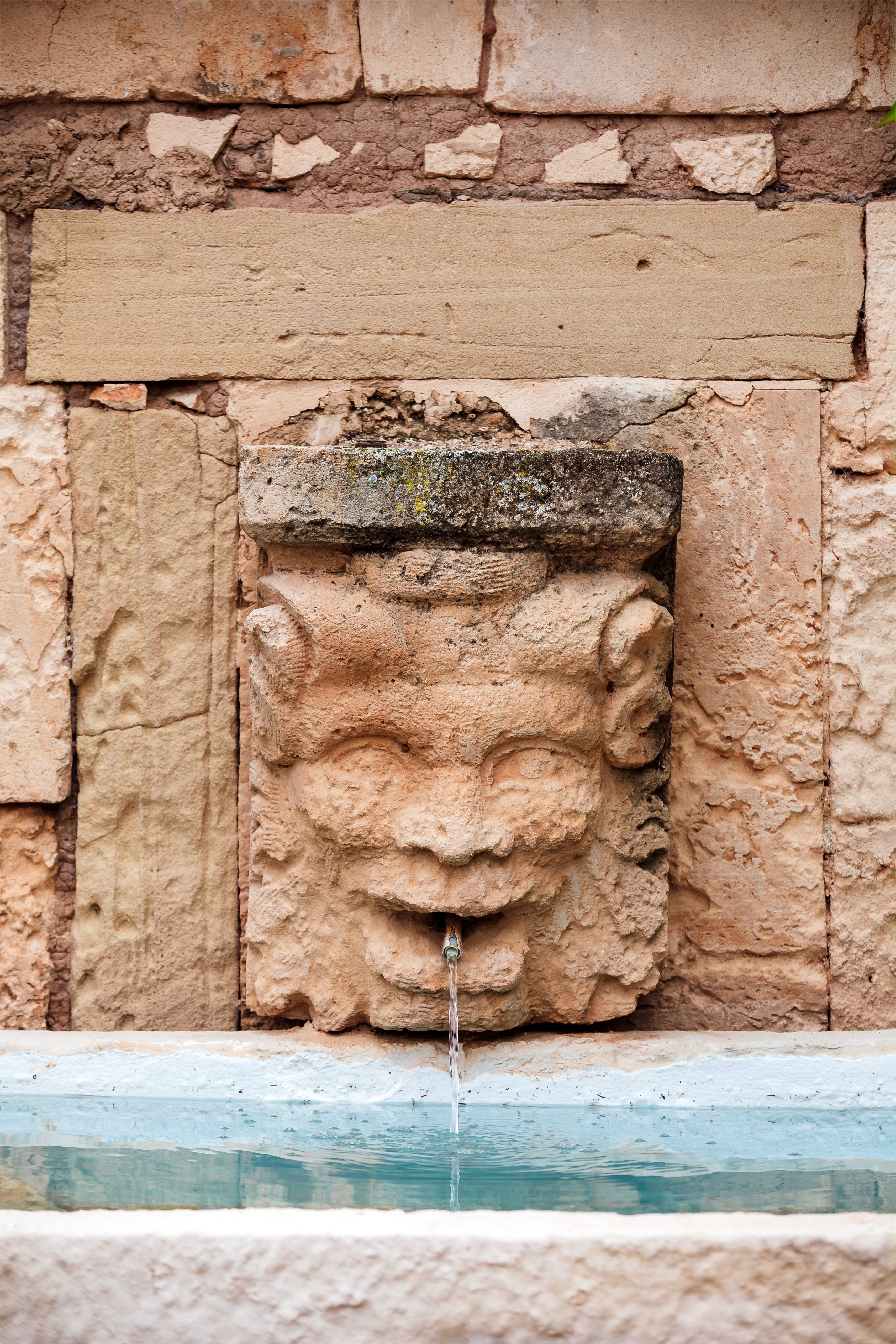 Weathered stone fountain at Cal Reiet, a carved face spouting water into a pale blue basin