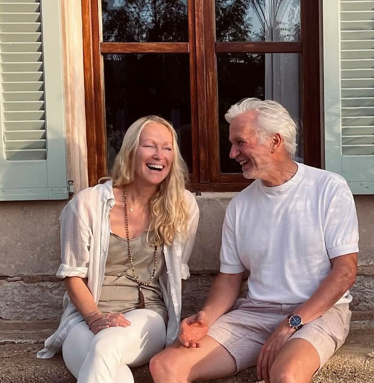 Henning Bensland laughing with a companion, seated together in front of a shuttered window at Cal Reiet