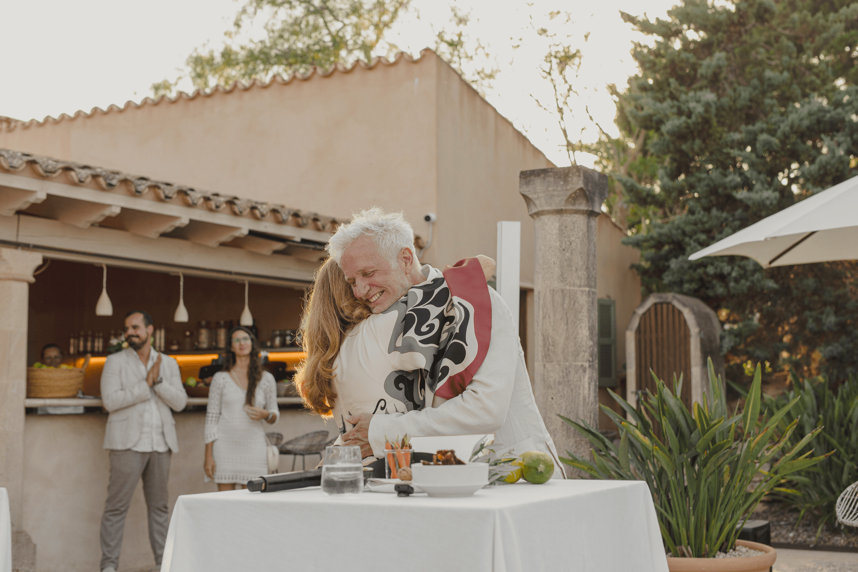 Henning Bensland embracing his wife Petra at a gathering in the courtyard of Cal Reiet, friends applauding by the garden bar behind them