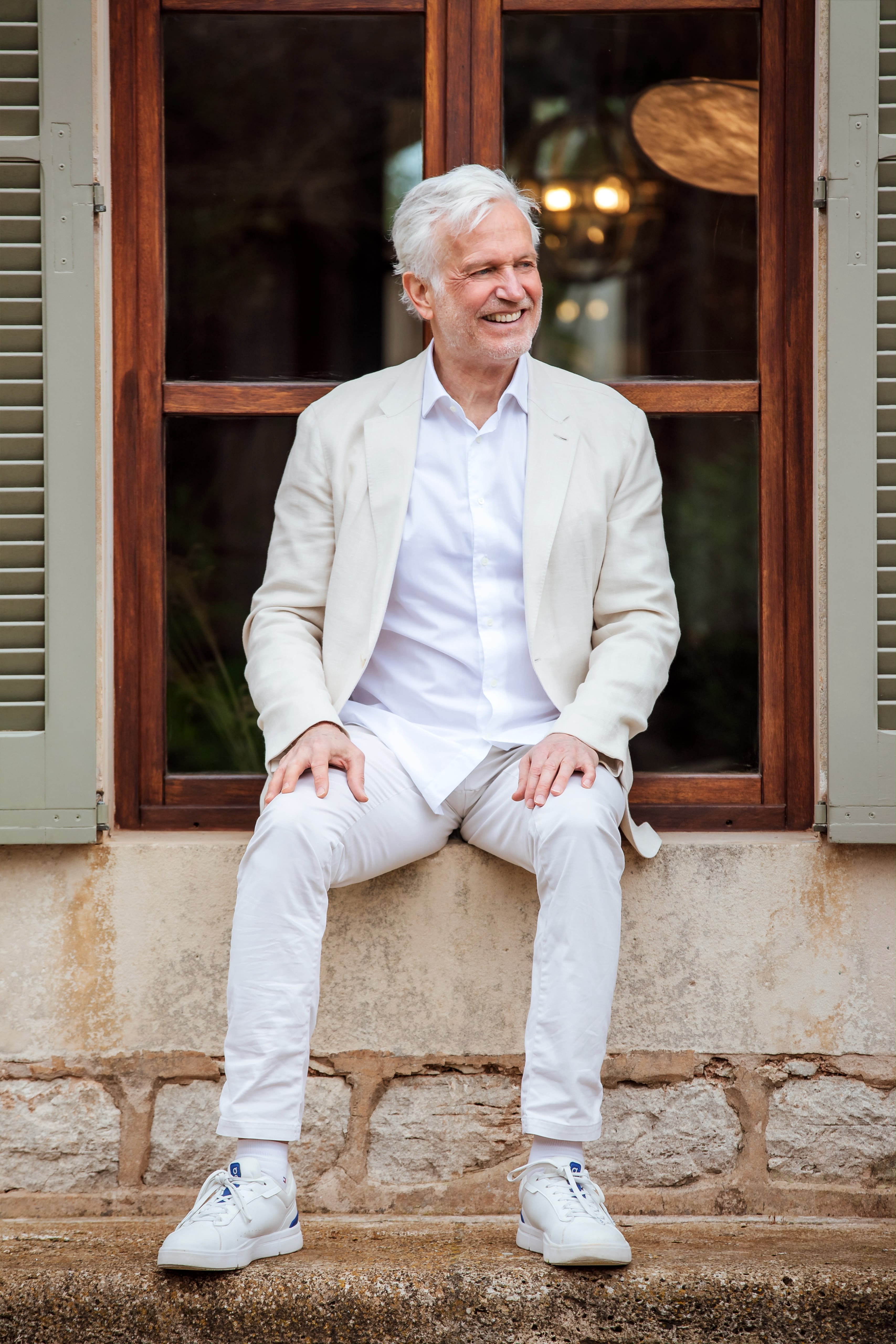 Henning Bensland in a cream linen suit, sitting on a stone windowsill in front of green-shuttered windows at Cal Reiet, smiling and looking to the side