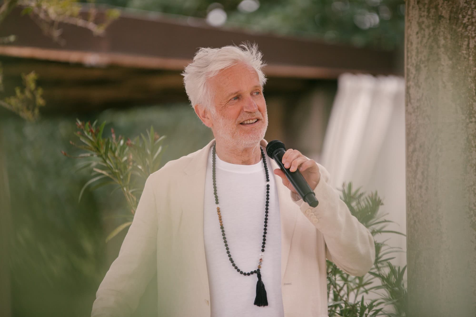 Henning Bensland speaking into a microphone outdoors, wearing a mala necklace and cream linen jacket