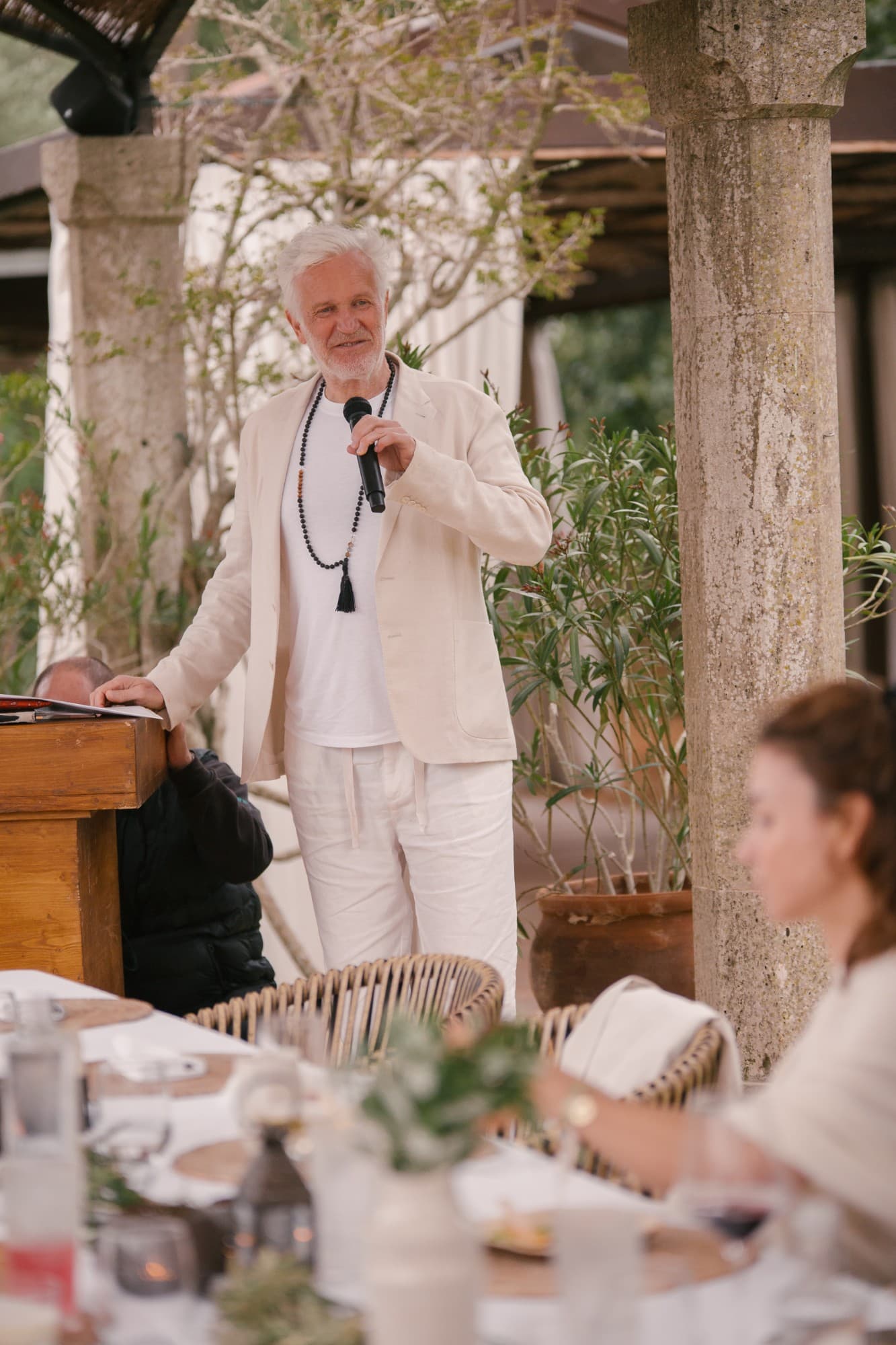 Henning Bensland speaking into a microphone at an outdoor podium, wearing a mala necklace and cream linen suit